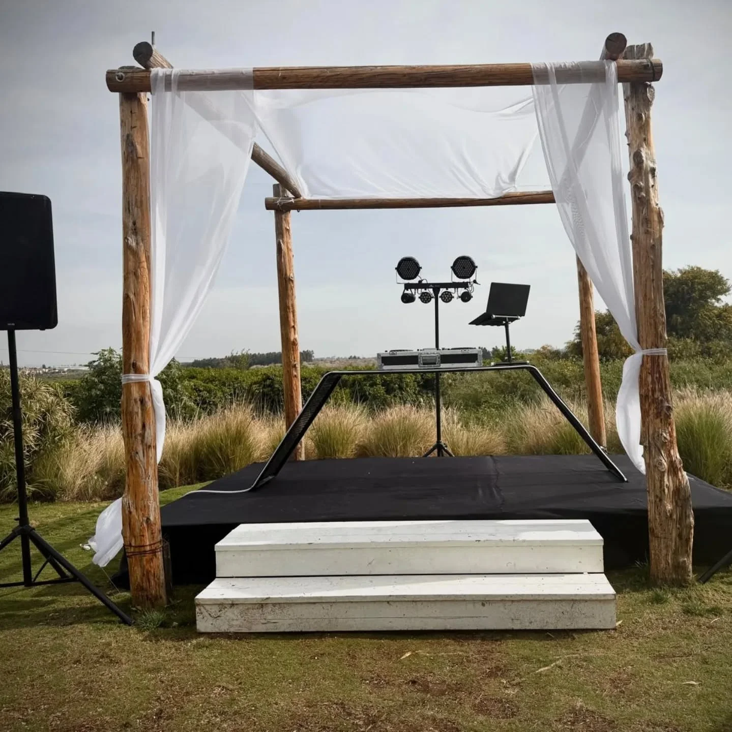 Outdoor DJ booth with speakers and laptop under a white draped wooden canopy.